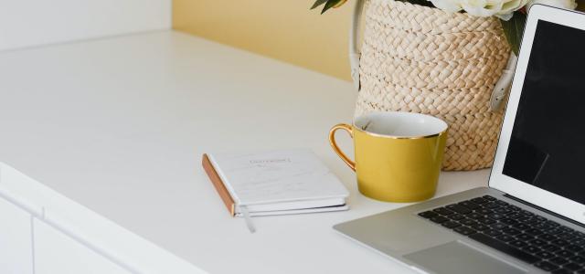 A desk with a laptop, a yellow coffee cup, and a notebook on it. Next to the laptop is a vase filled with white flowers